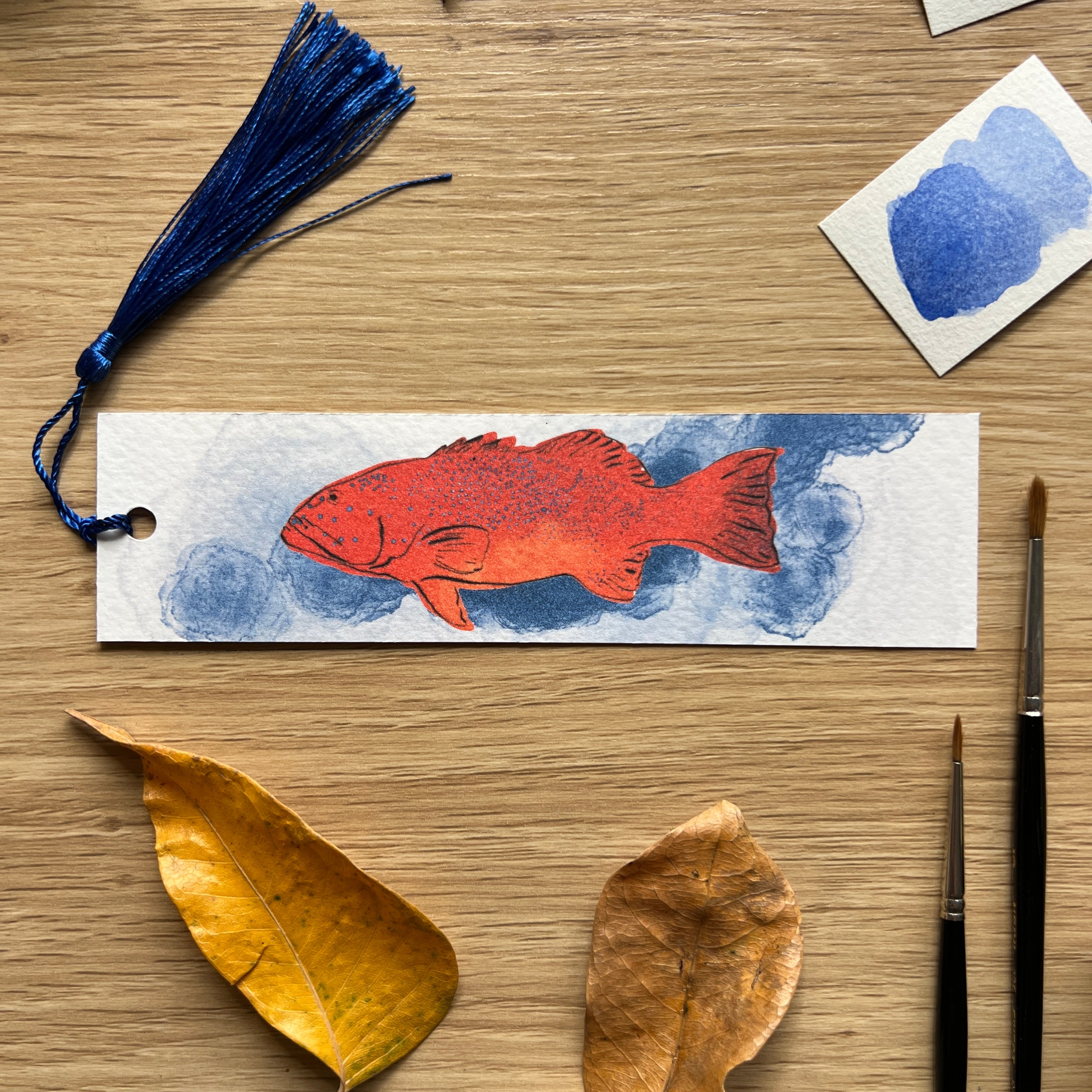 Bookmark with red fish illustration on a wooden surface with leaves and a paintbrush.