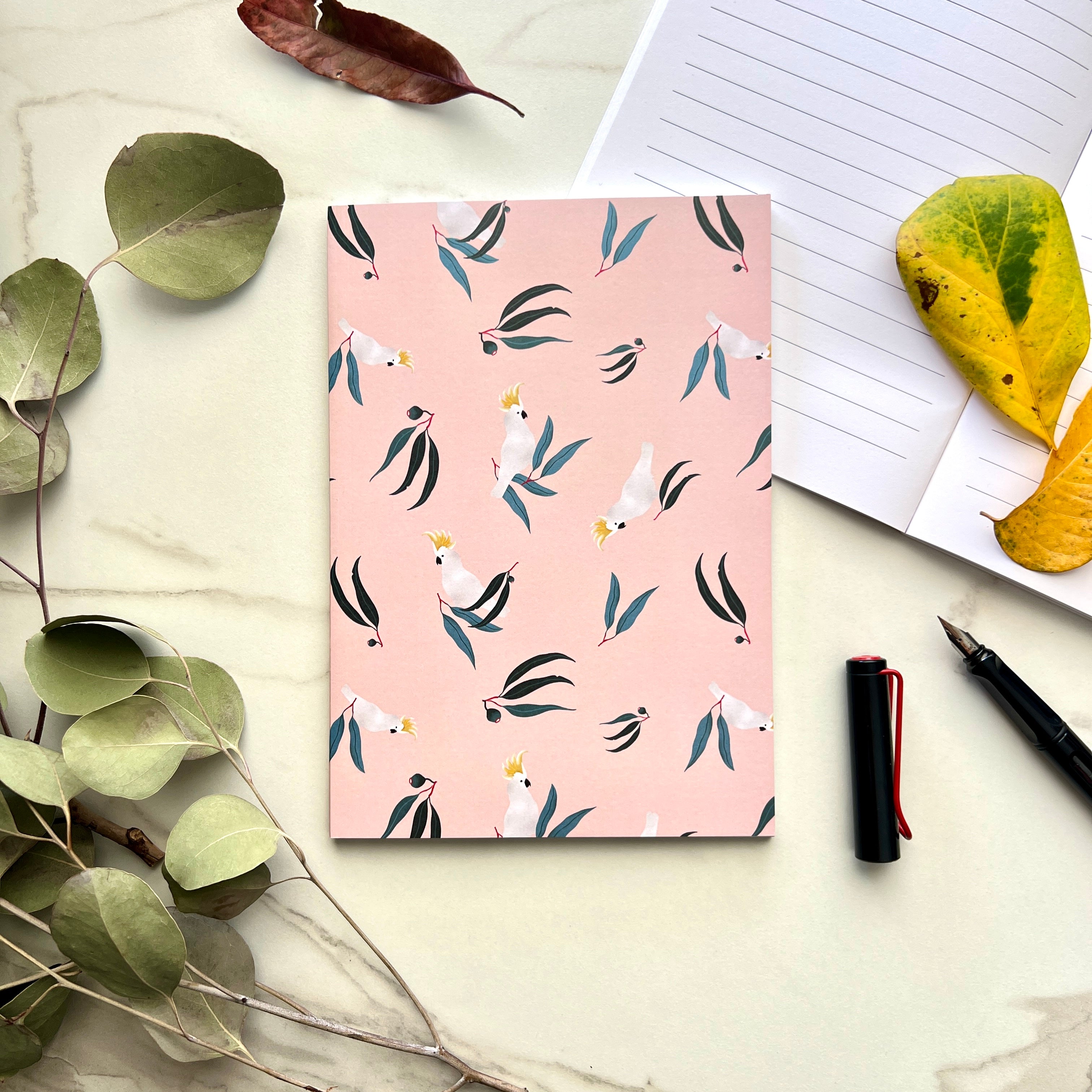 Notebook with a cockatoo and leaf pattern on a light surface with leaves and a pen.