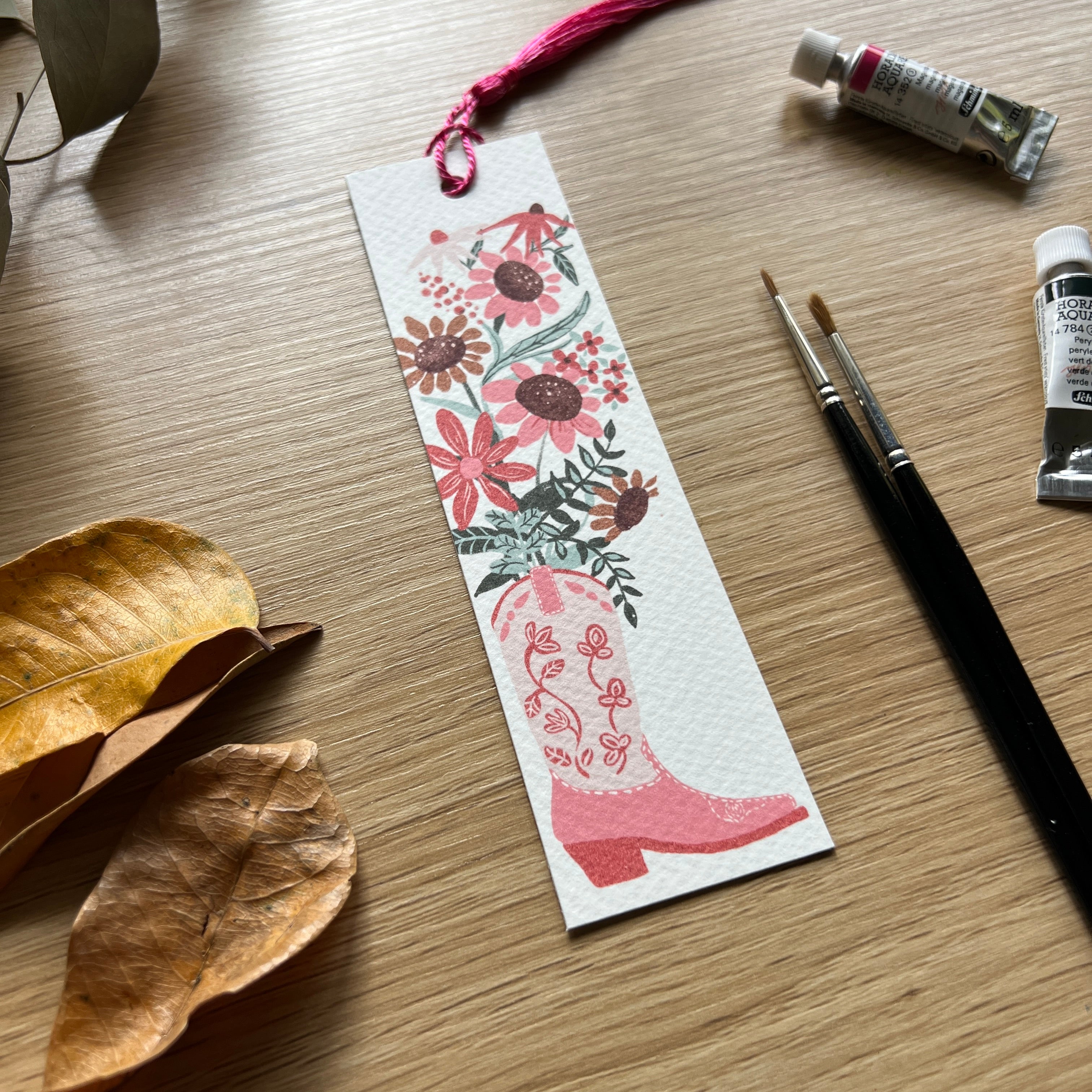 Floral bookmark with pink cowgirl boot design on a wooden surface with paint supplies.