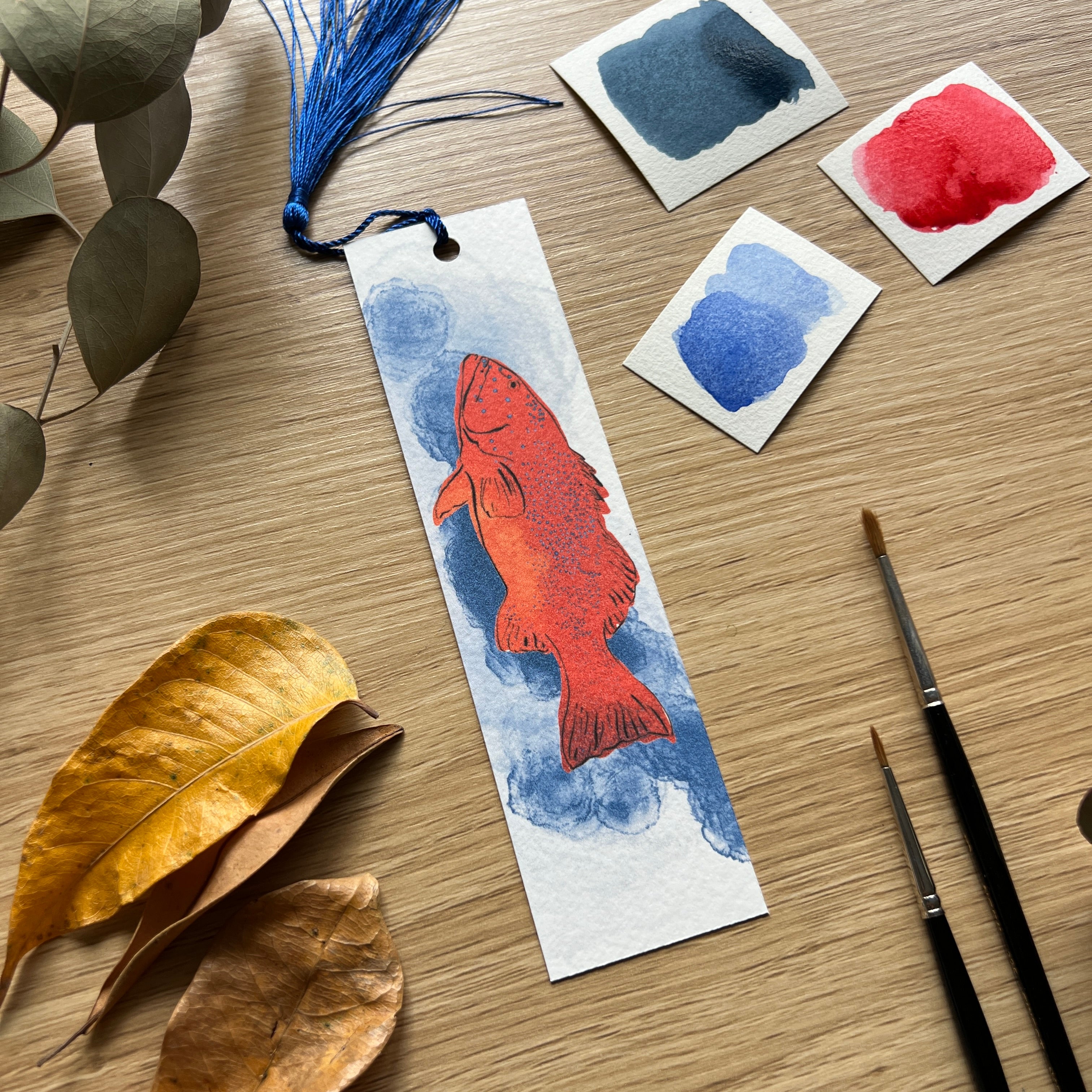 Handmade bookmark with red fish design on a wooden surface with watercolor samples and leaves.