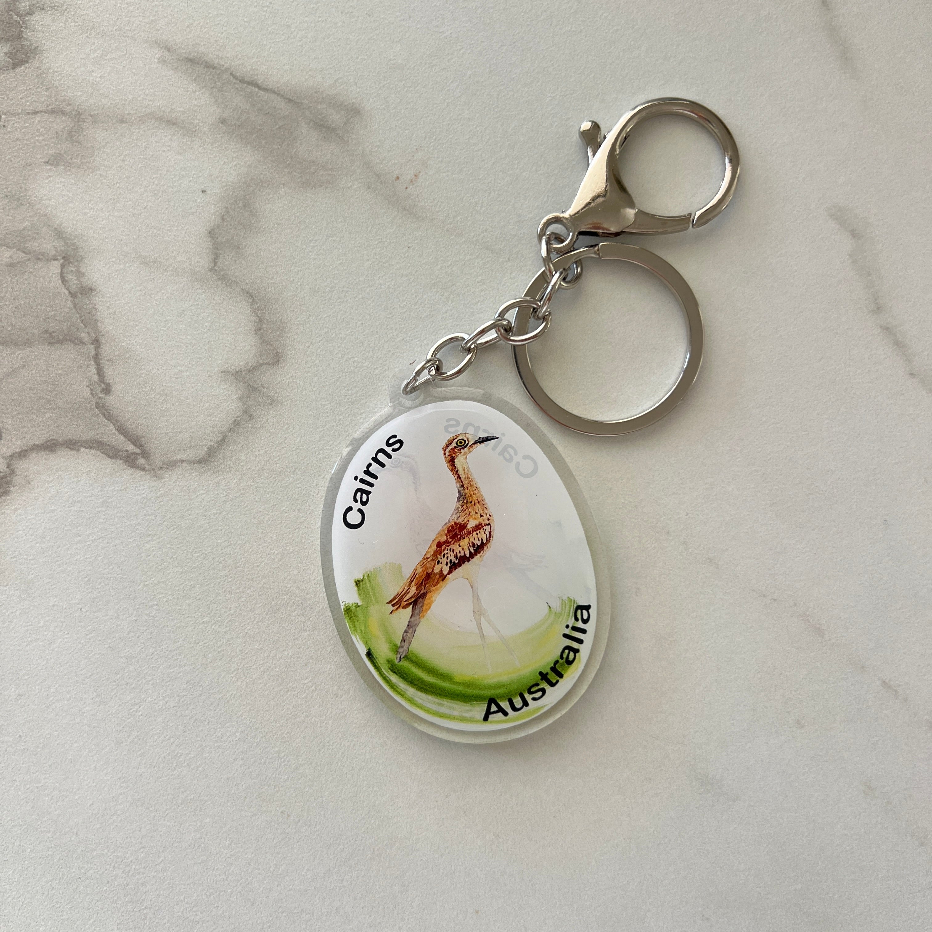 An oval acrylic keychain featuring a painted wombat illustration with the text 'Cairns 2026 Australia' on a silver clasp, displayed on a light gray marble surface.
