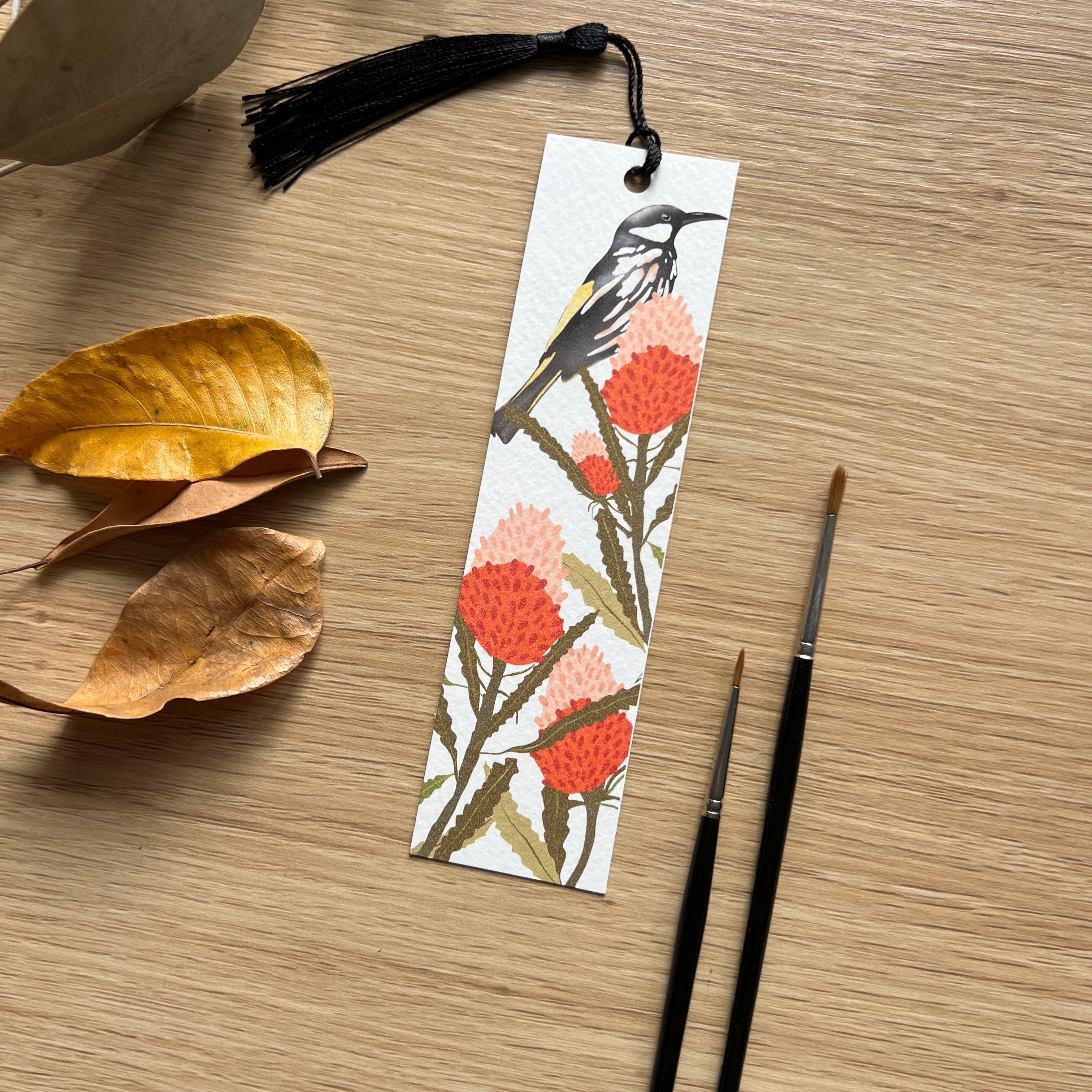 Bookmark with bird and floral design on a wooden surface with leaves and paintbrushes.