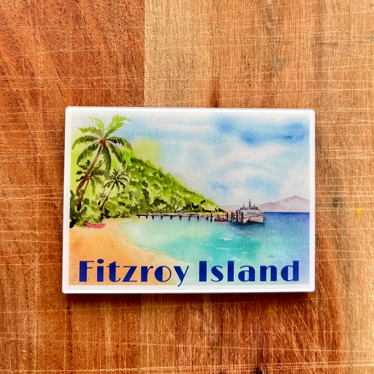 A rectangular souvenir magnet featuring a watercolor painting of Fitzroy Island, with palm trees, turquoise water, a pier, and a ferry, displayed on a wooden cutting board.