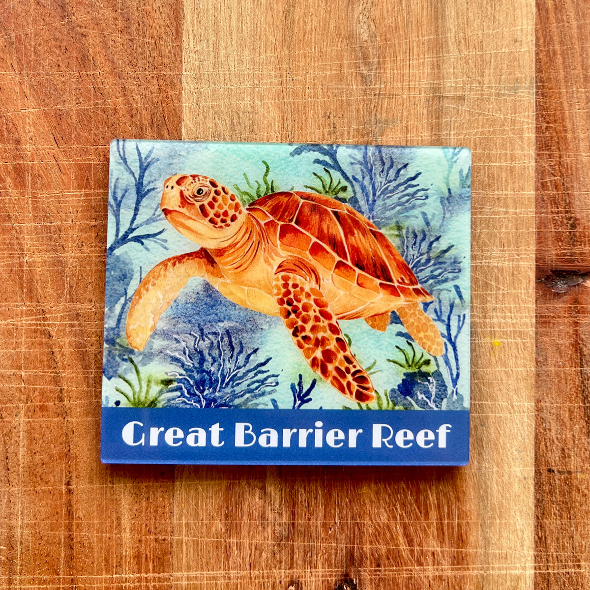 A square fridge magnet featuring a painted sea turtle on a coral reef with the text 'Great Barrier Reef' on a blue band at the bottom, placed on a wooden surface.