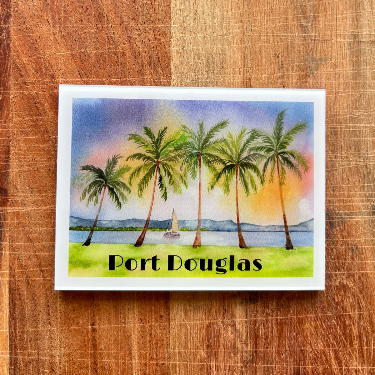 A rectangular fridge magnet featuring a watercolor tropical scene with palm trees, water, distant hills, and the text 'Port Douglas,' placed on a wooden surface.