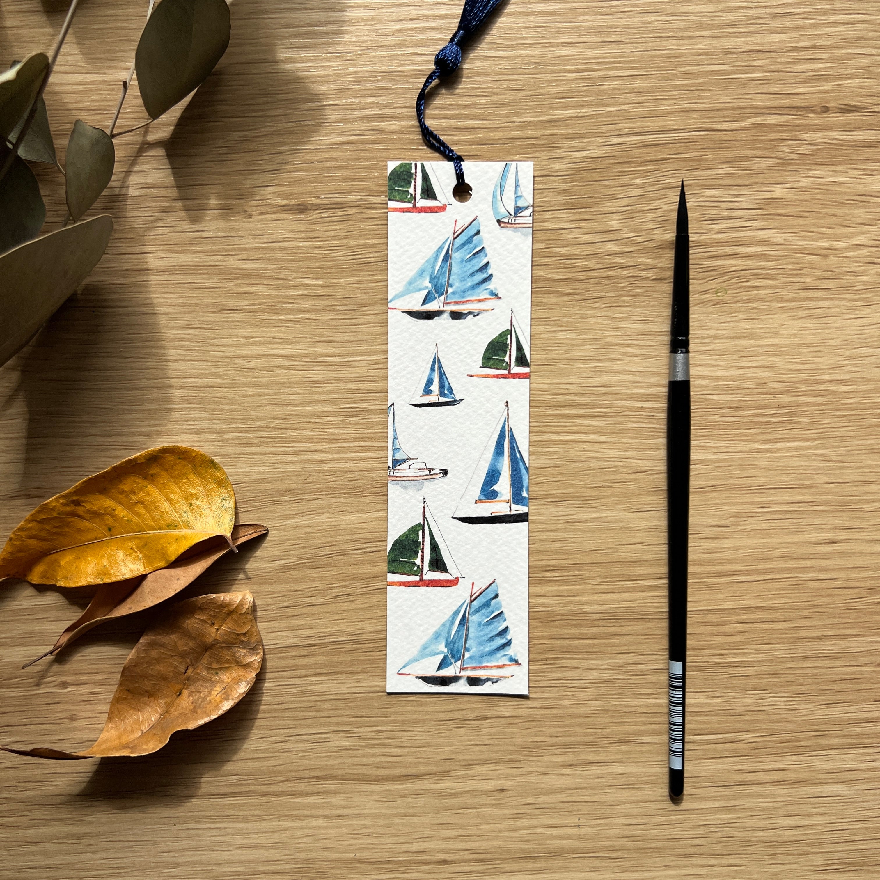 Bookmark with watercolour sailboat design next to a pen on a wooden surface with leaves.