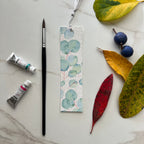 Bookmark with eucalyptus design, paintbrush, and paint tubes on a marble surface with leaves.
