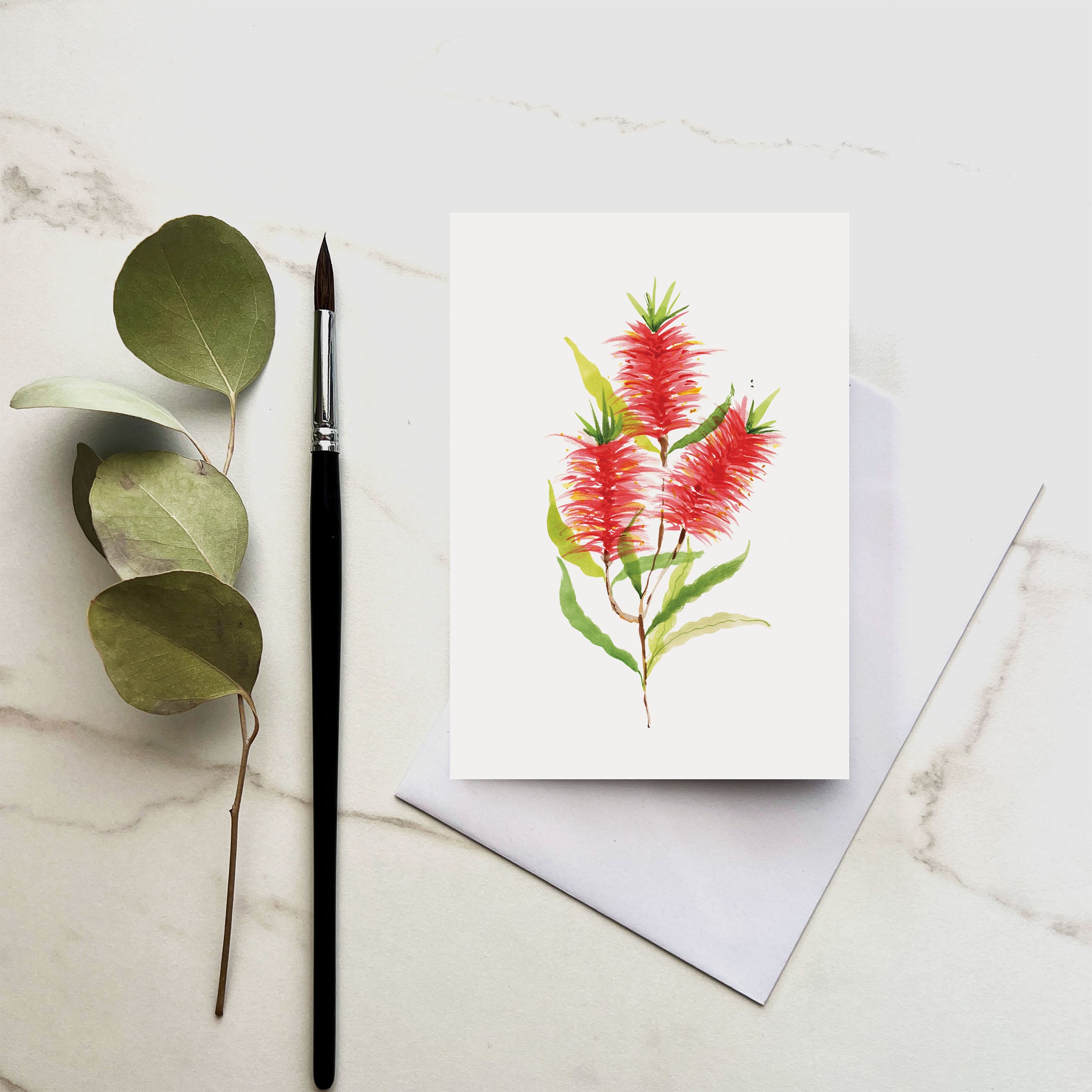 Greeting card with red flowers and green leaves on a marble surface with a paintbrush and leaves.