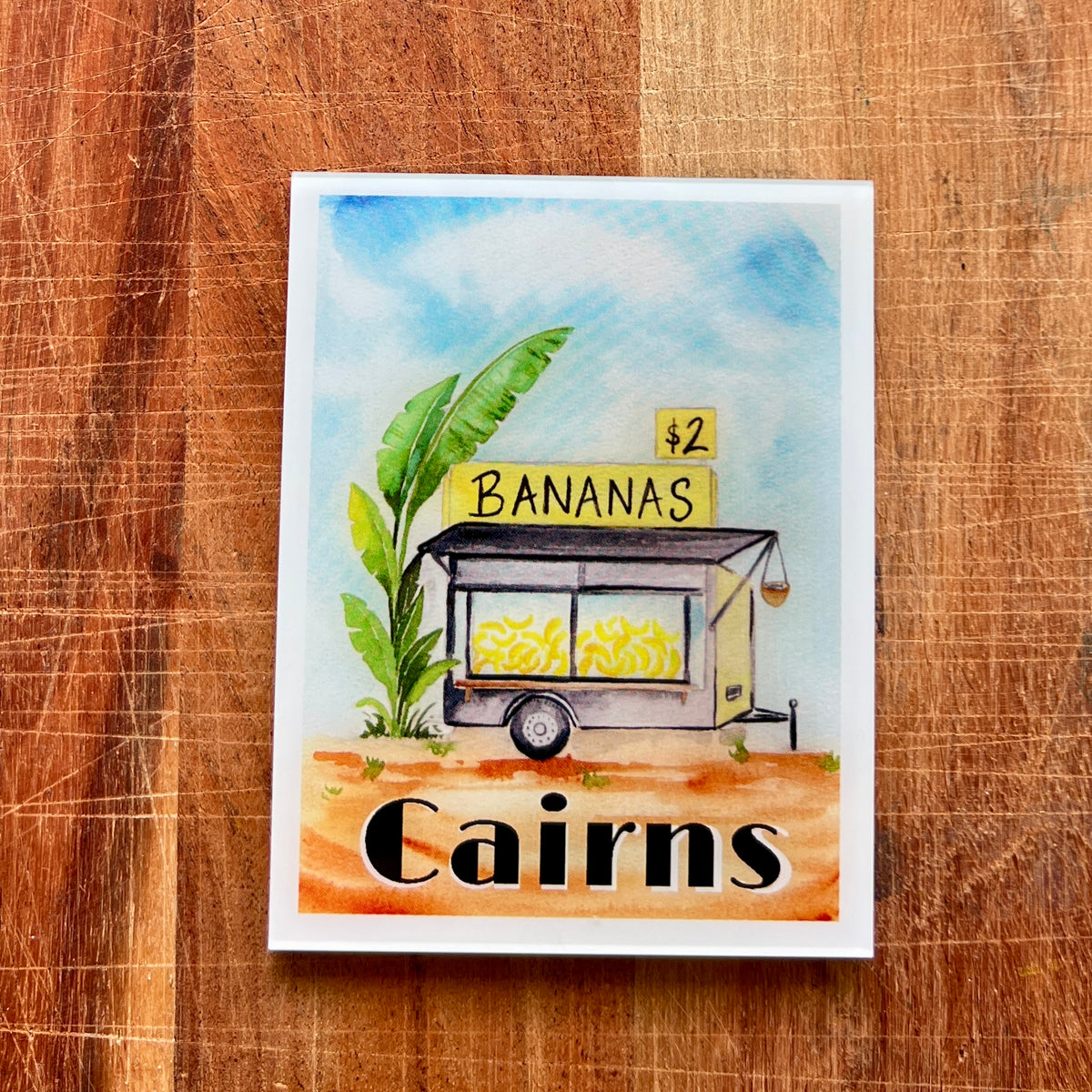 A square watercolor-style fridge magnet featuring a small banana stand with the words '$2 BANANAS' and 'Cairns' at the bottom, placed on a wooden surface.