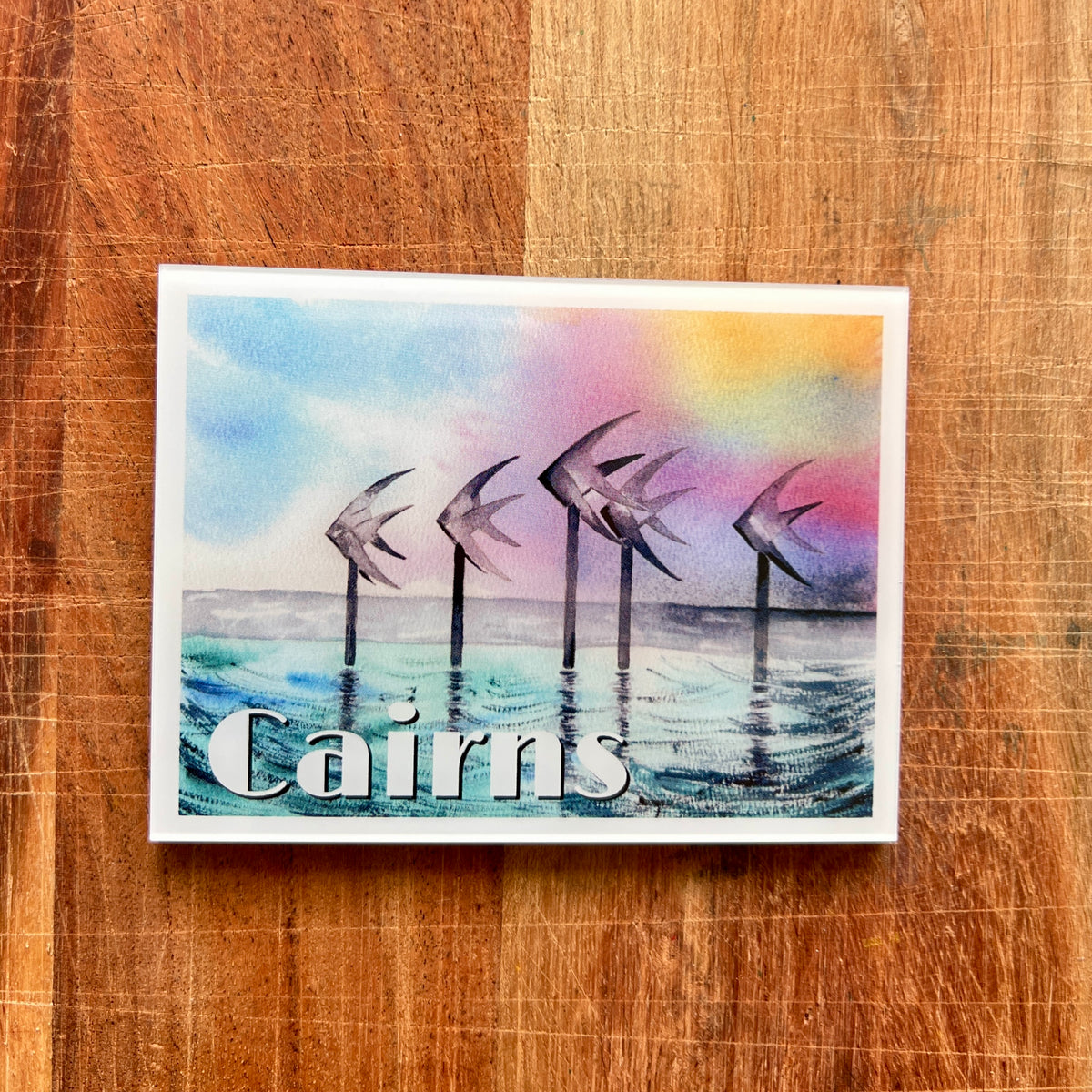 A rectangular fridge magnet with a watercolor-style coastal scene showing several gulls over blue water and the word "Cairns" at the bottom, placed on a wooden surface.
