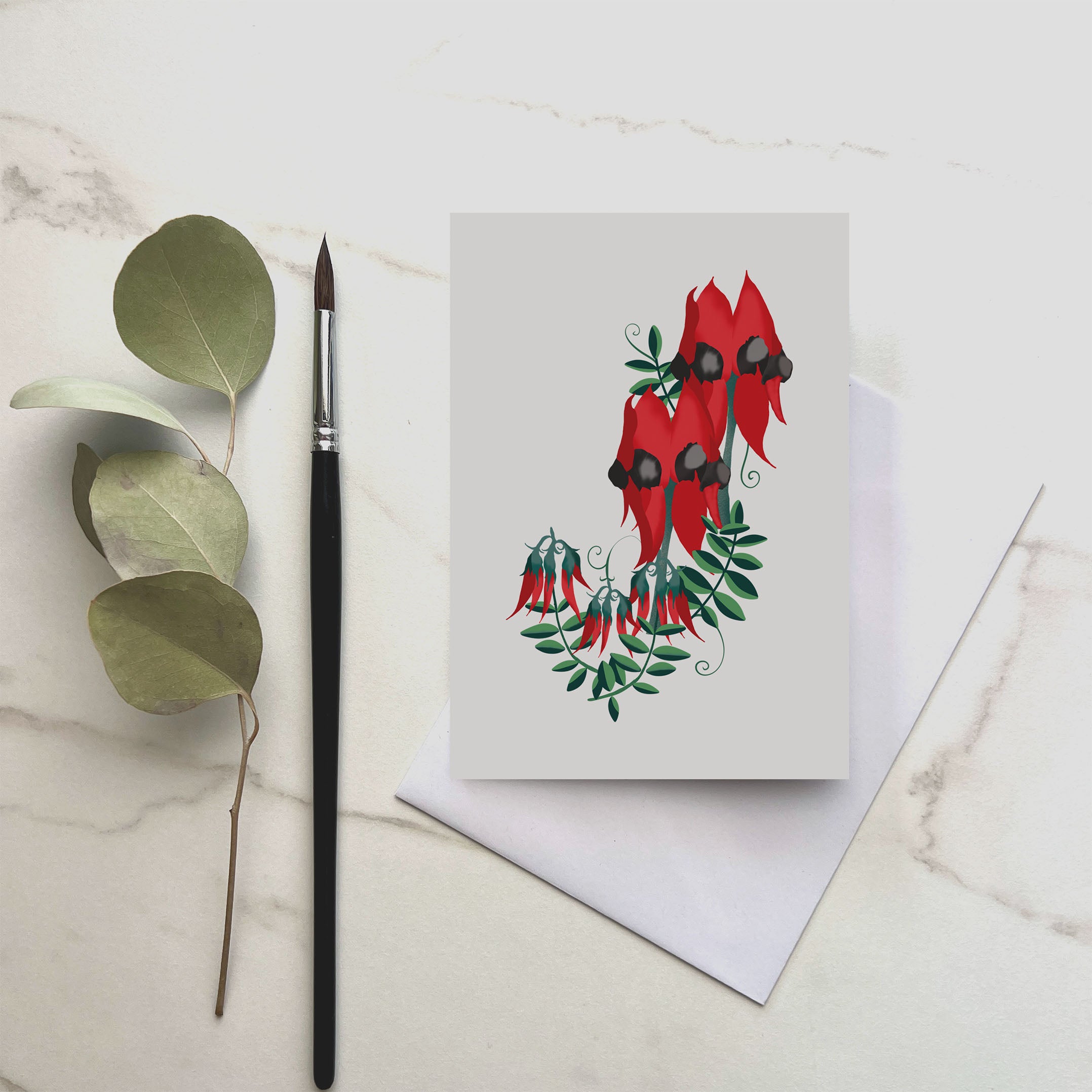 Greeting card with red floral design, brush, and eucalyptus leaves on marble surface