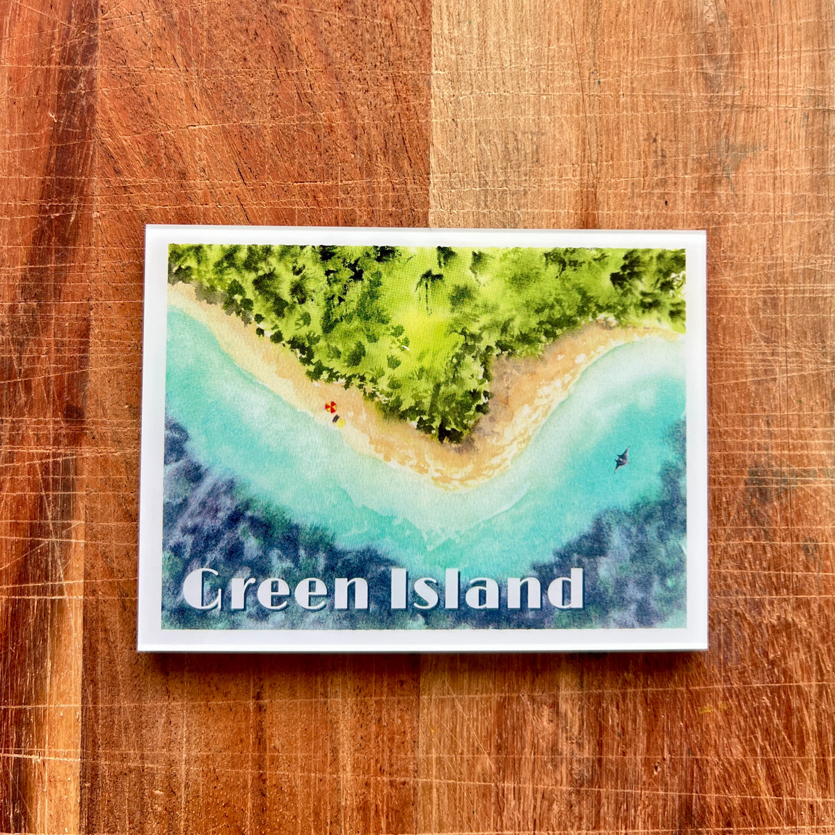 A rectangular art print or postcard featuring a watercolor-style aerial view of Green Island, with turquoise water, sandy beach, and green forest, placed on a wooden surface. The words 'Green Island' are printed across the lower part of the artwork.