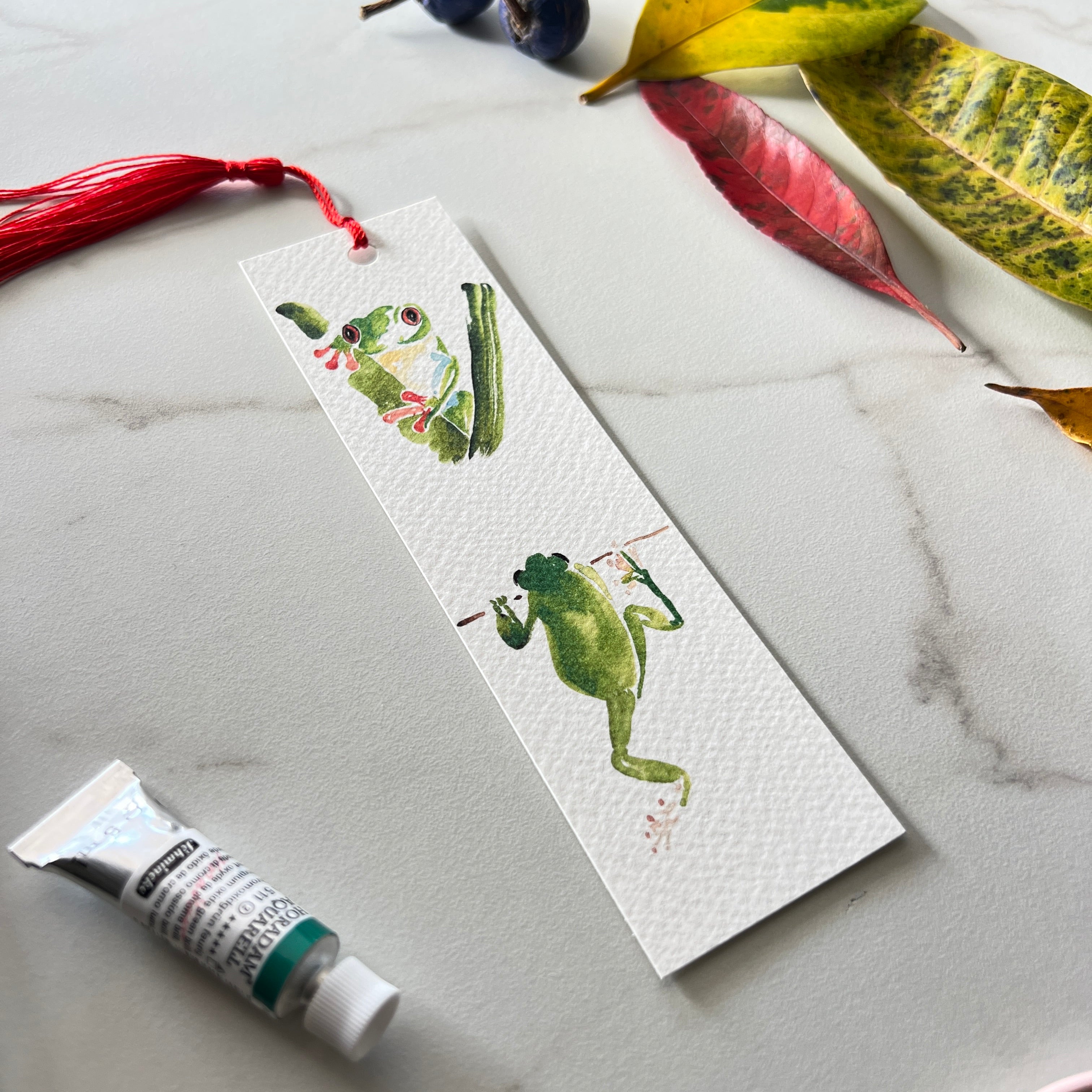 Bookmark with frog illustration on a marble surface with leaves and a tube of paint.