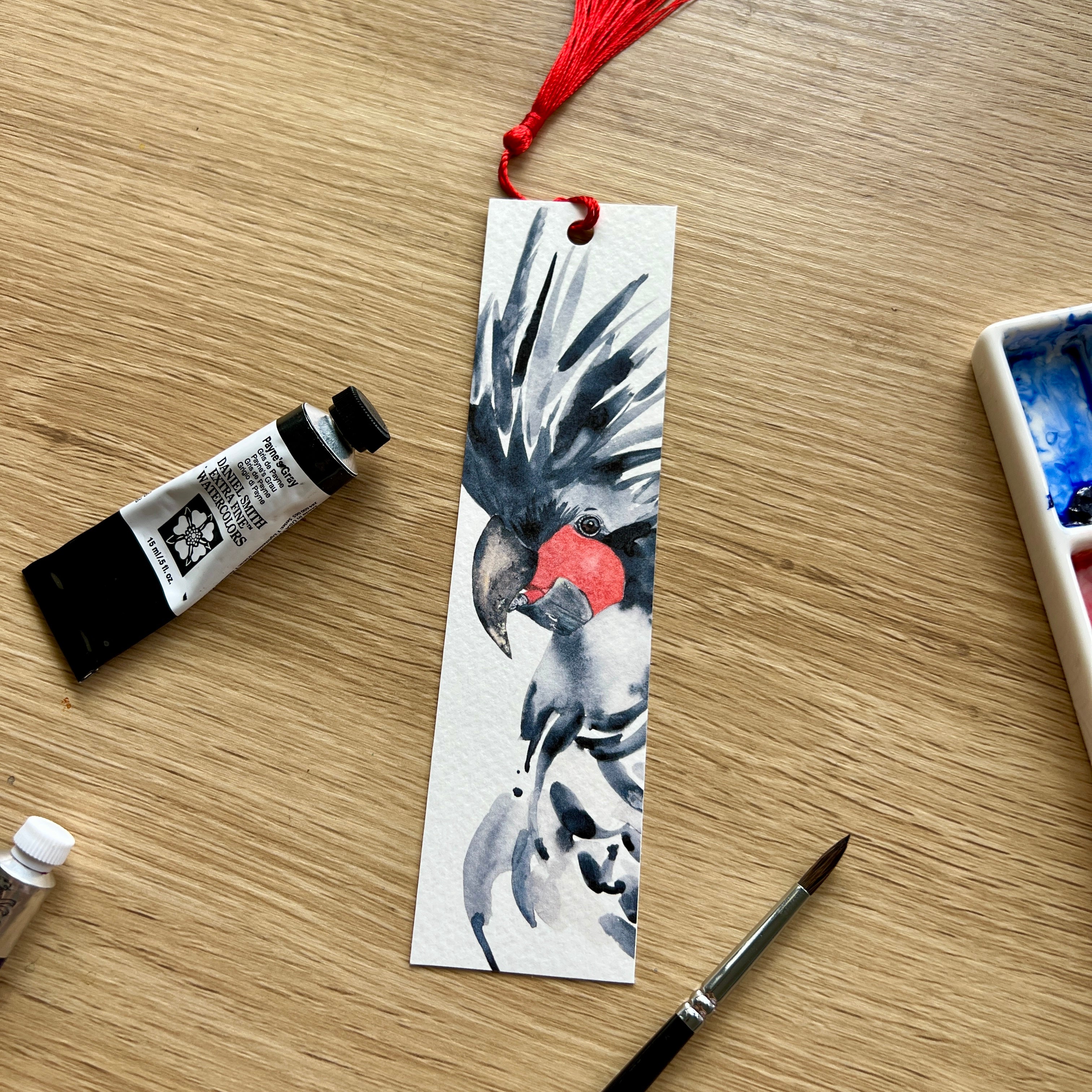 watercolour bookmark with a palm cockatoo design on a wooden surface with painting supplies.