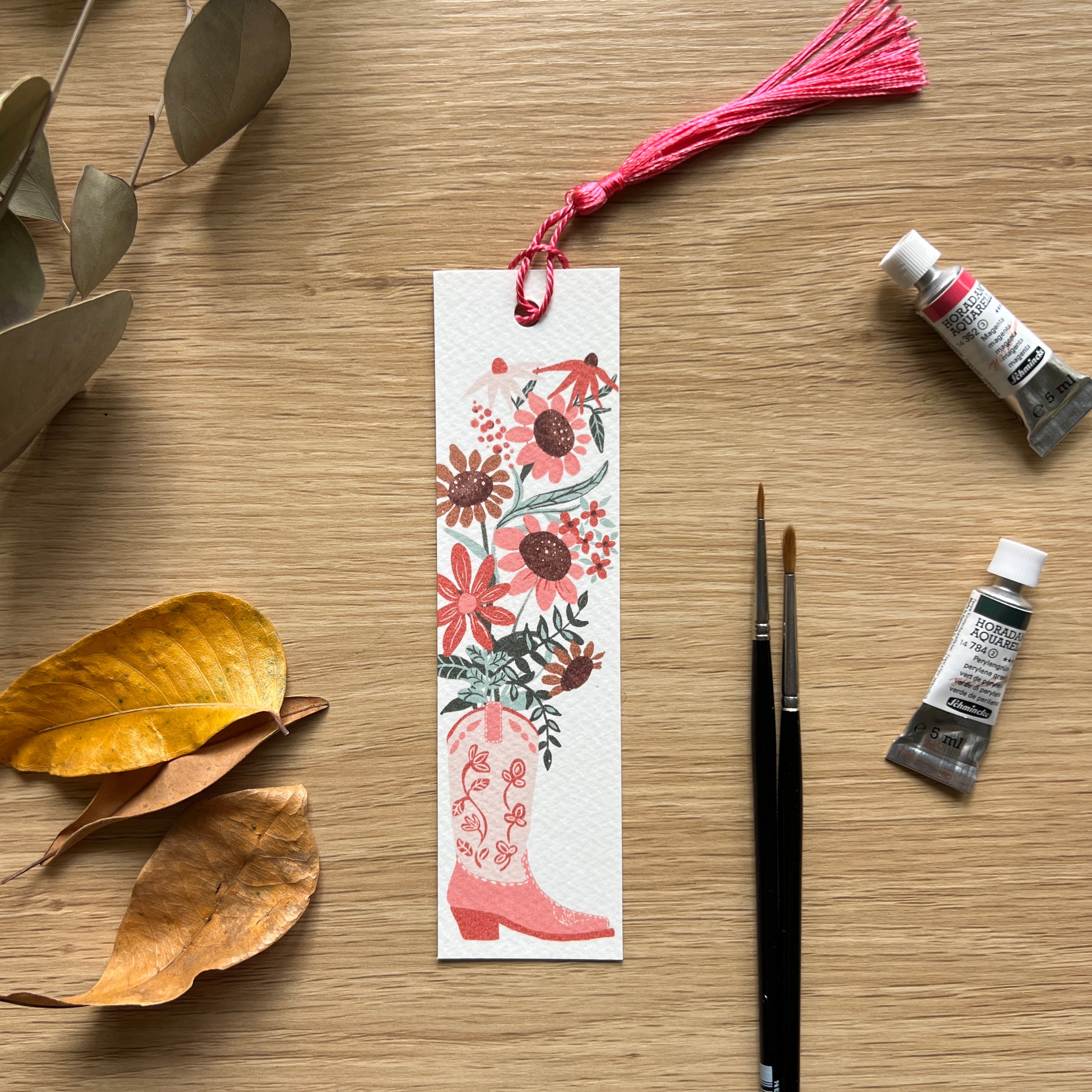 Decorative bookmark with floral design and pink cowboy boot on a wooden surface with paint tubes and brushes.