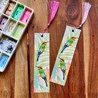 Two colorful rainbow bee eater bookmarks on a wooden surface with an art palette and tassels