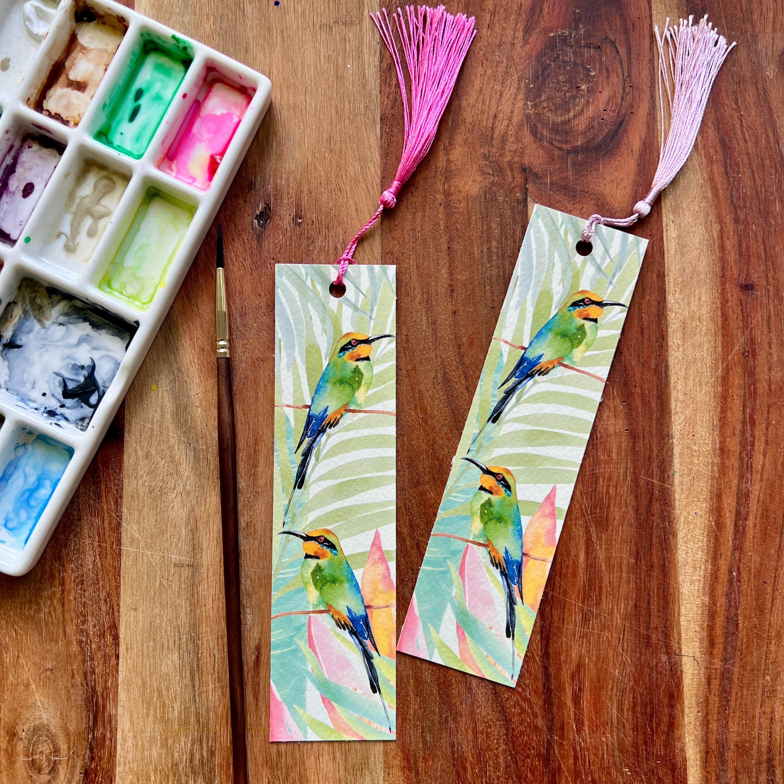 Two colorful rainbow bee eater bookmarks on a wooden surface with an art palette and tassels