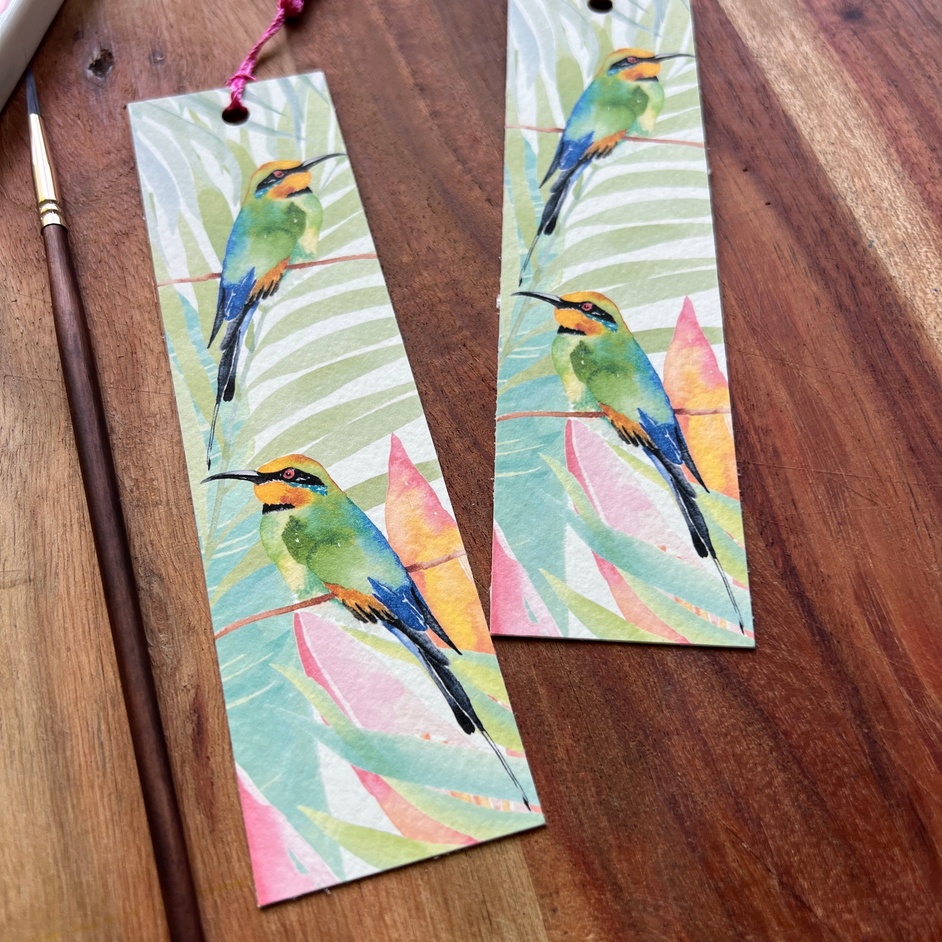 Two colorful bird bookmarks on a wooden surface with a paintbrush.