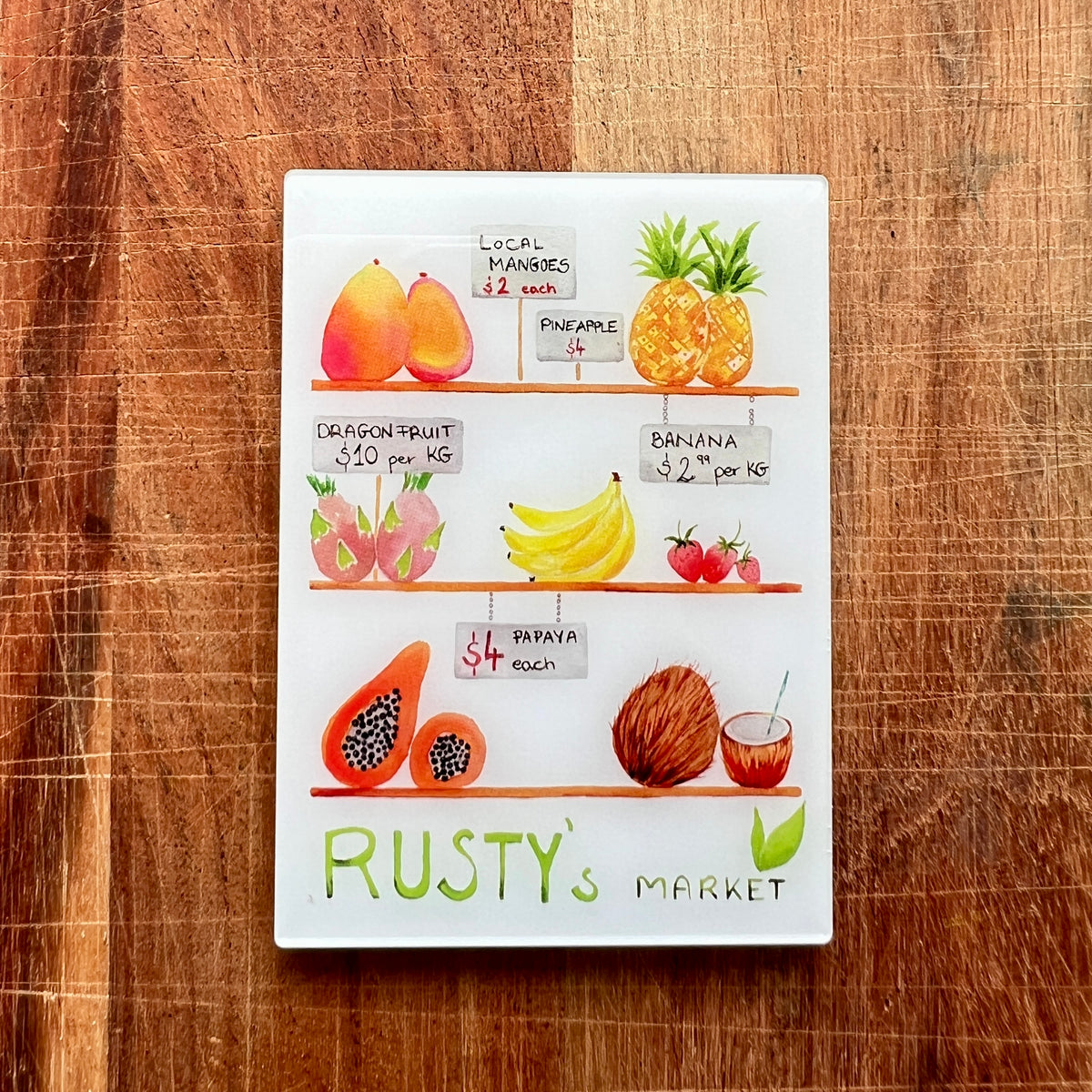 A rectangular fridge magnet featuring a colorful watercolor-style illustration of fruits on shelves with handwritten prices, signed 'RUSTY'S MARKET' at the bottom, placed on a rustic wooden surface.