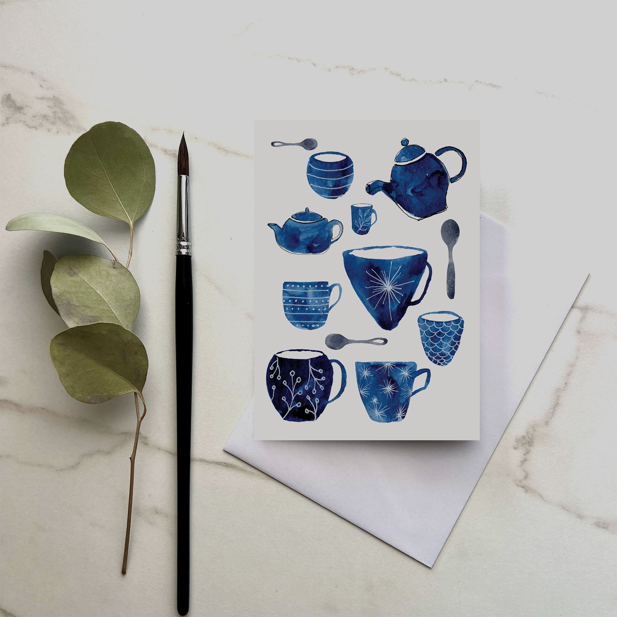 Card with blue teacup design on a marble surface with a brush and leaves.