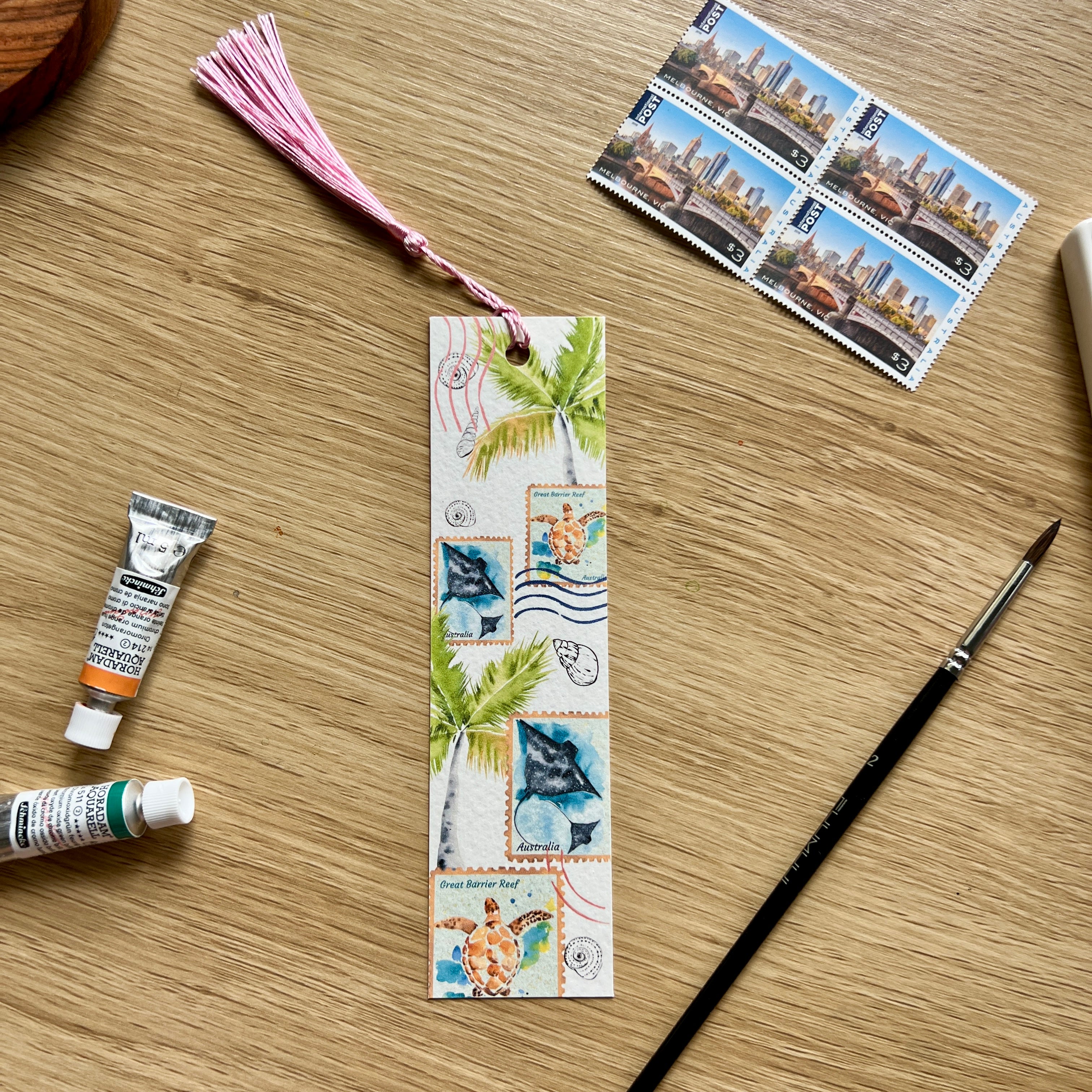 Bookmark with tropical design, paint tubes, and stamps on a wooden surface