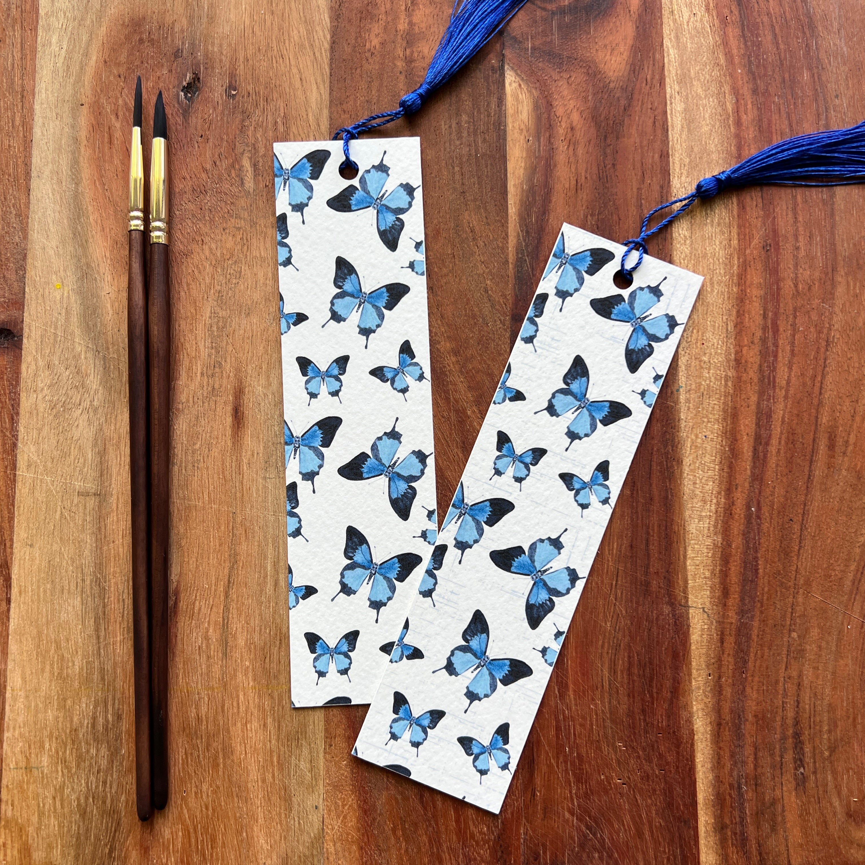 Two bookmarks with blue butterfly pattern on a wooden surface with a paintbrush and thread.
