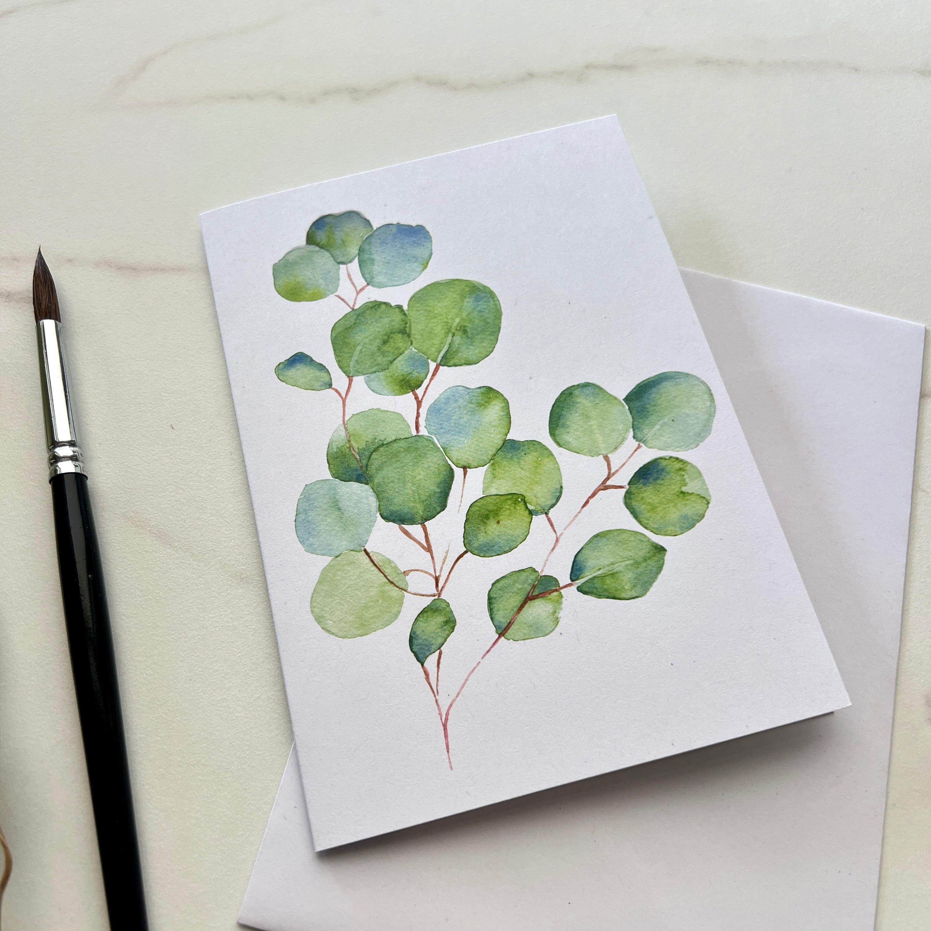 Greeting card with eucalyptus design on a white surface with a paintbrush.