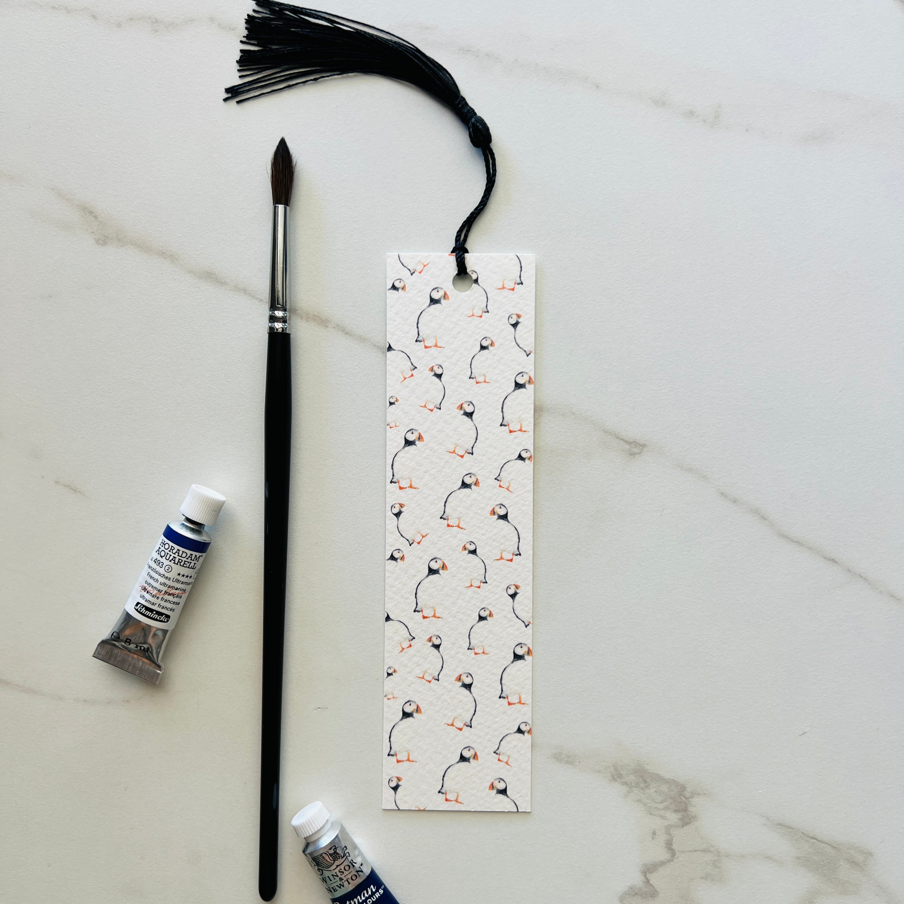 watercolour puffin bookmark with tassel on light background with paint brush and paint tubes 