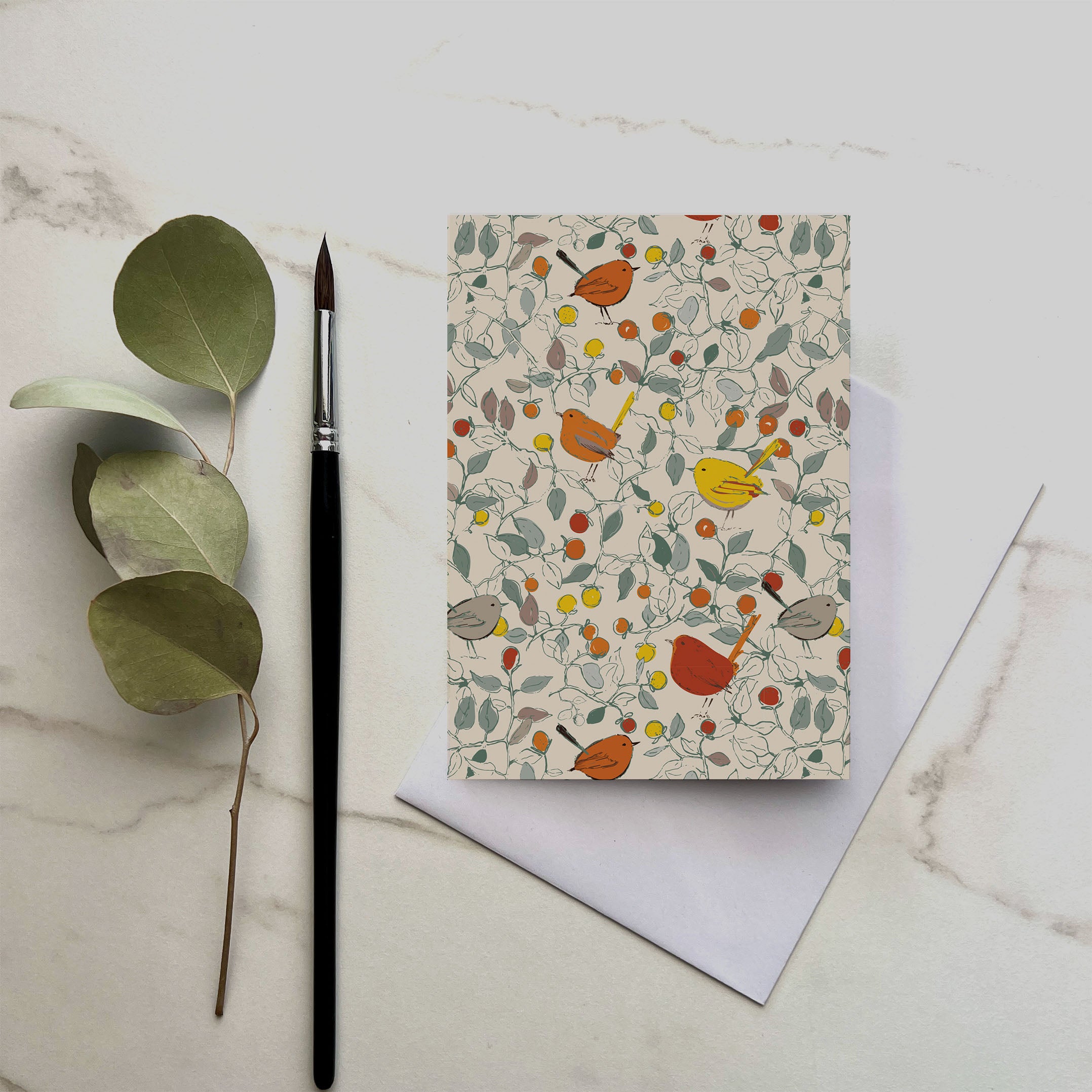 Greeting card with bird and floral pattern on a marble surface with a brush and eucalyptus leaves.